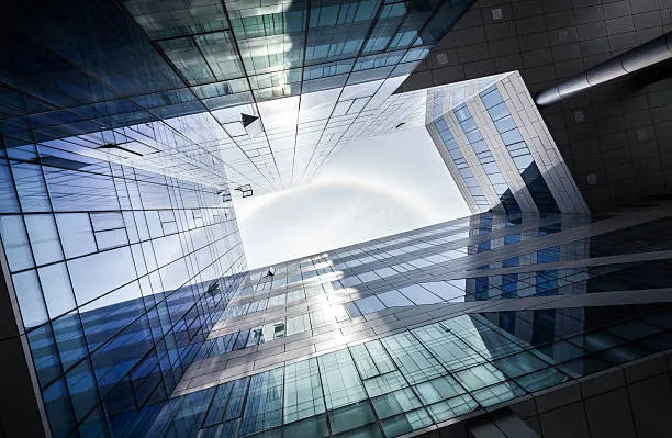 Looking up at the modern glass architecture of a building, showcasing sharp lines, reflective surfaces, and geometric patterns against a bright sky.